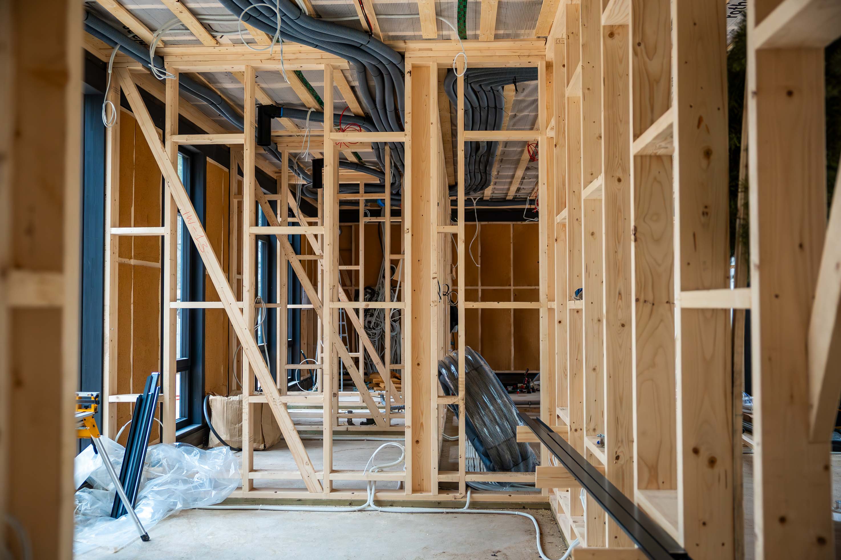 Interior view of a house under construction featuring wooden framing, exposed ducts, electrical wiring, large windows, and scattered construction materials.